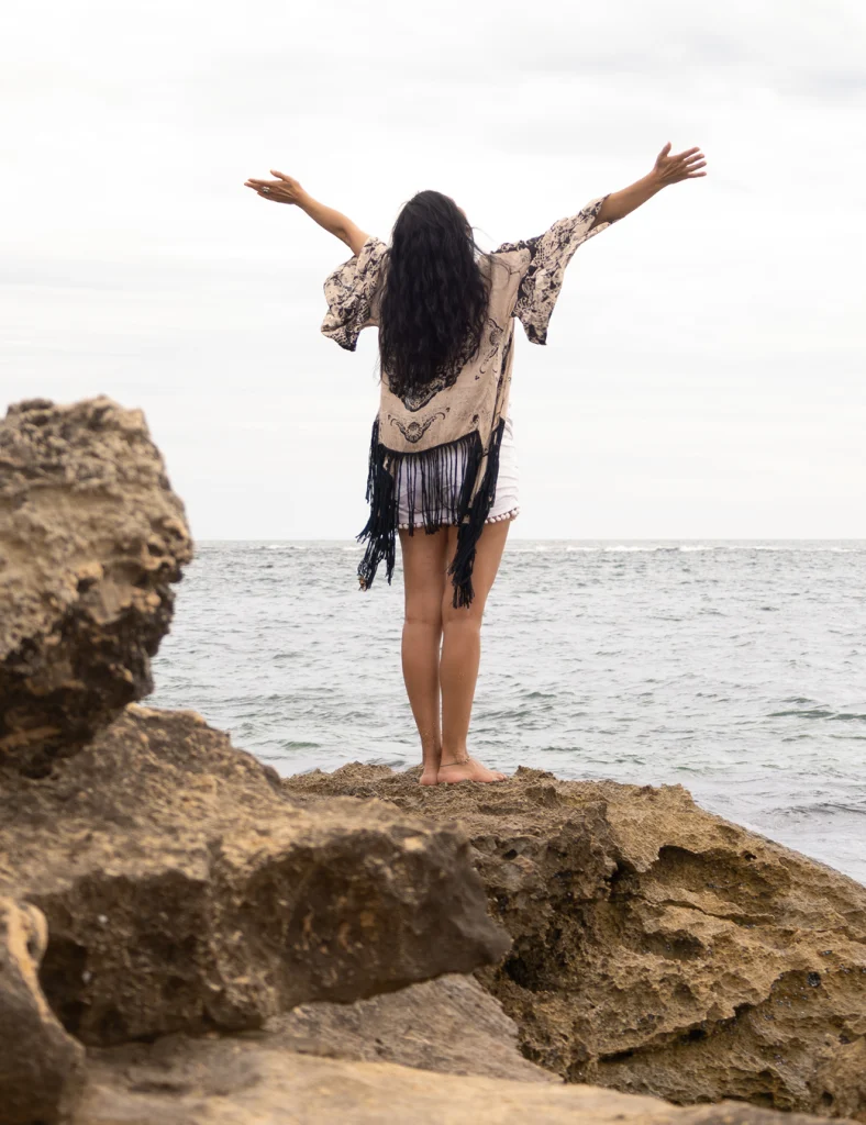 Veronica Galipo, life coach and self-love guide, standing with arms raised in the open air at the beach.